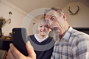 Grandson With Grandfather Pulling Faces And Taking Selfie On Mobile Phone At Home