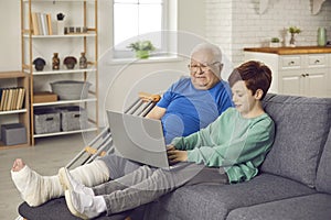 Grandson and his grandpa with broken leg sitting on sofa and using laptop together