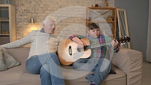 The grandson plays for his grandfather guitar, music. An elderly man is sitting on with a young fat guy playing guitar