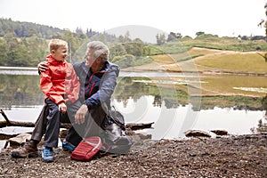 Grandson sitting on his grandfather`s knee at the shore of a lake smiling, Lake District, UK