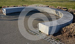 A granite park bench in a horseshoe shaped arch of light stone around a lawn and flower beds with ornamental grasses.