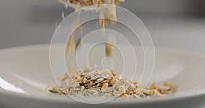 Granola with almond flakes and pumpkin seeds fall into white bowl closeup