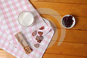 Granola bar, glass of milk, dried strawberry and a bowl of dry cranberry on a towel on wooden table flat lay