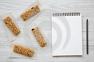 Granola bars on baking sheet and blank notepad over white wooden background, top view.