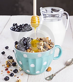 Granola with blueberry, mint, honey and milk in blue bowl with milk jug on a white table and metal spoon