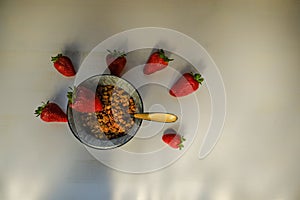 Granola in bowl with spoon across ripe strawberries on white wooden table top view. Healthy organic breakfast. Copy space