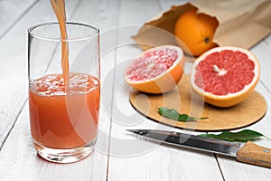 Grapefruit juice is poured into a glass on a wooden table, fresh cut oranges in the background
