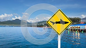 Graphic of shark on yellow sign on blurred sea and restaurant in background to warn people not to swim due to sharks