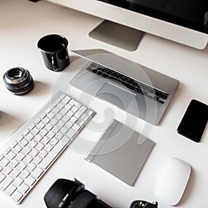 Graphics tablet, camera, laptop, black cup, objects, keyboard, mobile phone and wireless mouse on a white wooden table.