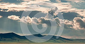 Grass hills on horizon in steppe under heavy clouds sky during sunset with sun light beams.