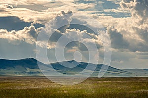 Grass hills on horizon in steppe under heavy clouds sky during sunset with sun light beams.