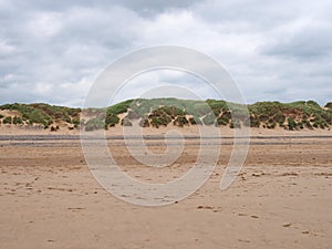 Grassy sand dunes at the edge of a flat sandy beach with footprints in summer sunlight with blue sky and clouds