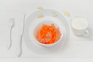 Grated fresh carrots in a plate. On the table is a knife and fork, garlic cloves. Sour cream sauce. White background.