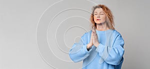 Grateful hopeful middle age woman stands in meditative pose, enjoys peaceful atmosphere, holds hands in praying gesture, has sense