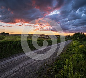 Gravel countryside road through spring meadow, cloudy evening sunset sky, rural hills, village and fields in far