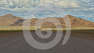 Gravel road with lonely tree covered by cloud shadow and sun shining on hills and slightly green meadows near Solitaire, Namibia.