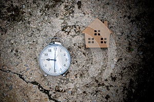 A gray alarm clock and small wooden house toy on cement floor.