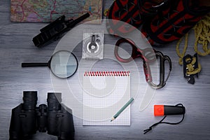 On a gray background, is a set of traveler comfortable shoes, rope, carabiners, magnifying glass, binoculars, compass, map, walkie