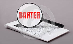 On a gray background, a white calculator and a magnifying glass with the text BARTER. Business concept