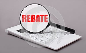 On a gray background, a white calculator and a magnifying glass with the text REBATE. Business concept