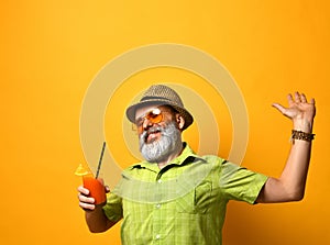 Gray-bearded man in hat, green shirt, sunglasses. Smiling, stretching himself, holding glass of juice, posing on orange background