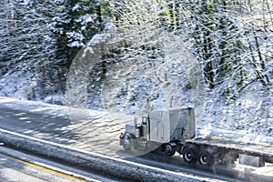 Gray big rig powerful classic American semi truck with empty flat bed semi trailer running on winter snow road to pick up the
