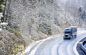 Gray big rig semi truck with covered semi trailer transporting cargo on winter snowing road with wet surface
