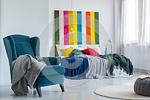 Gray blanket on a comfy, blue armchair next to a colorful bed with cushions in a bright bedroom interior with striped painting on