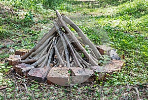 Gray and brown firewood for a bonfire are in a round fire with orange bricks on green grass in a summer forest. like a pyramid