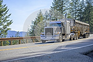 Gray classic big rig semi truck with vertical pipes transporting cargo in covered bulk semi trailer running on the mountain road