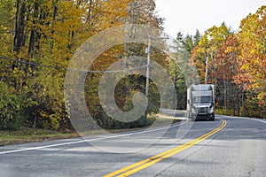 Gray commercial big rig semi truck with semi trailer running on the narrow winding road in autumn forest
