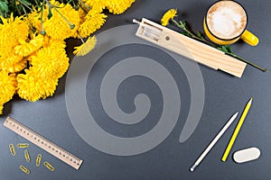 Gray desk surface with color pencils, eraser, ruler, wooden pencil box, big cup of cappuccino and bunch of yellow chrysanthemums.