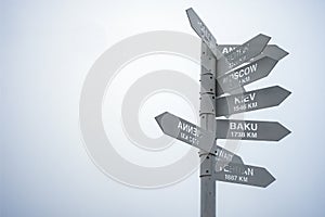 Gray direction sign pointing on capitals of countries on a gray foggy sky background.