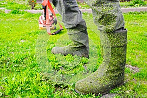 Gray dirty boots in the fresh green cut grass on the feet of the worker. Red trimmer in the background