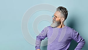 Gray-haired, bearded elderly man in sunglasses, purple sweater. Hands on hips, looking aside, posing on blue background. Close up