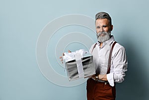 Gray-haired man in white shirt, brown pants and suspenders. Smiling, holding silver gift box, posing against blue background