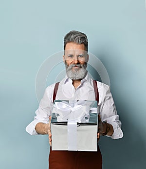 Gray-haired man in white shirt, brown pants and suspenders. Smiling, holding silver gift box, posing against blue background