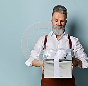 Gray-haired man in white shirt, brown pants and suspenders. Smiling, holding silver gift box, posing against blue background