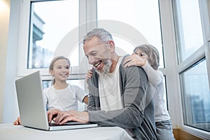 Gray-haired bearded man working on laptop while his kids interfering him