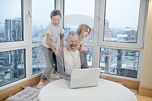 Gray-haired bearded man working on laptop while his kids interfering him