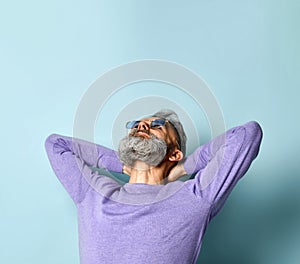 Gray-haired man in purple sweater, sunglasses. Put his hands behind head and looking up, posing on blue background. Close up