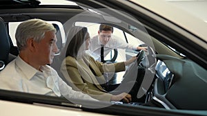 A gray-haired Caucasian woman is sitting behind the wheel of a car. Car dealership salesman consulting mature married