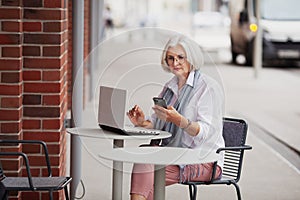 Gray haired elderly businesswoman sitting in cafe on street with mobile phone and laptop, senior woman working using