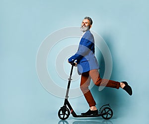 Gray-haired elderly man in white shirt, jacket, brown pants, loafers. Riding black scooter and posing sideways on blue background