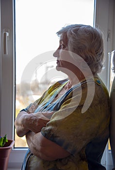 A gray-haired elderly woman stands at the window with her arms crossed over her chest