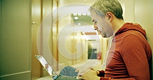 Gray haired man in casual clothes works on his laptop in the hotel hallway