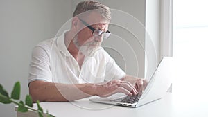 A gray-haired man with glasses and a beard works with a laptop sitting at a desk in the office. Work on removal