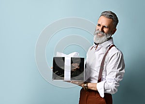 Gray-haired man in white shirt, brown pants and suspenders. Smiling, holding silver gift box, posing against blue background