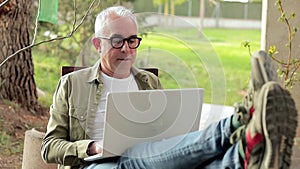 Gray-haired middle aged man using laptop outdoors. Man put feet on table and relax during a break