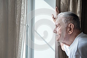 Gray-haired old man, forced to remain in quarantine due to the covid-19 coronavirus, looks out the window with a disconsolate